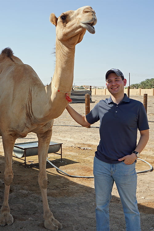 Harrin standing beside a dromedary camel with a green oasis in the distance.